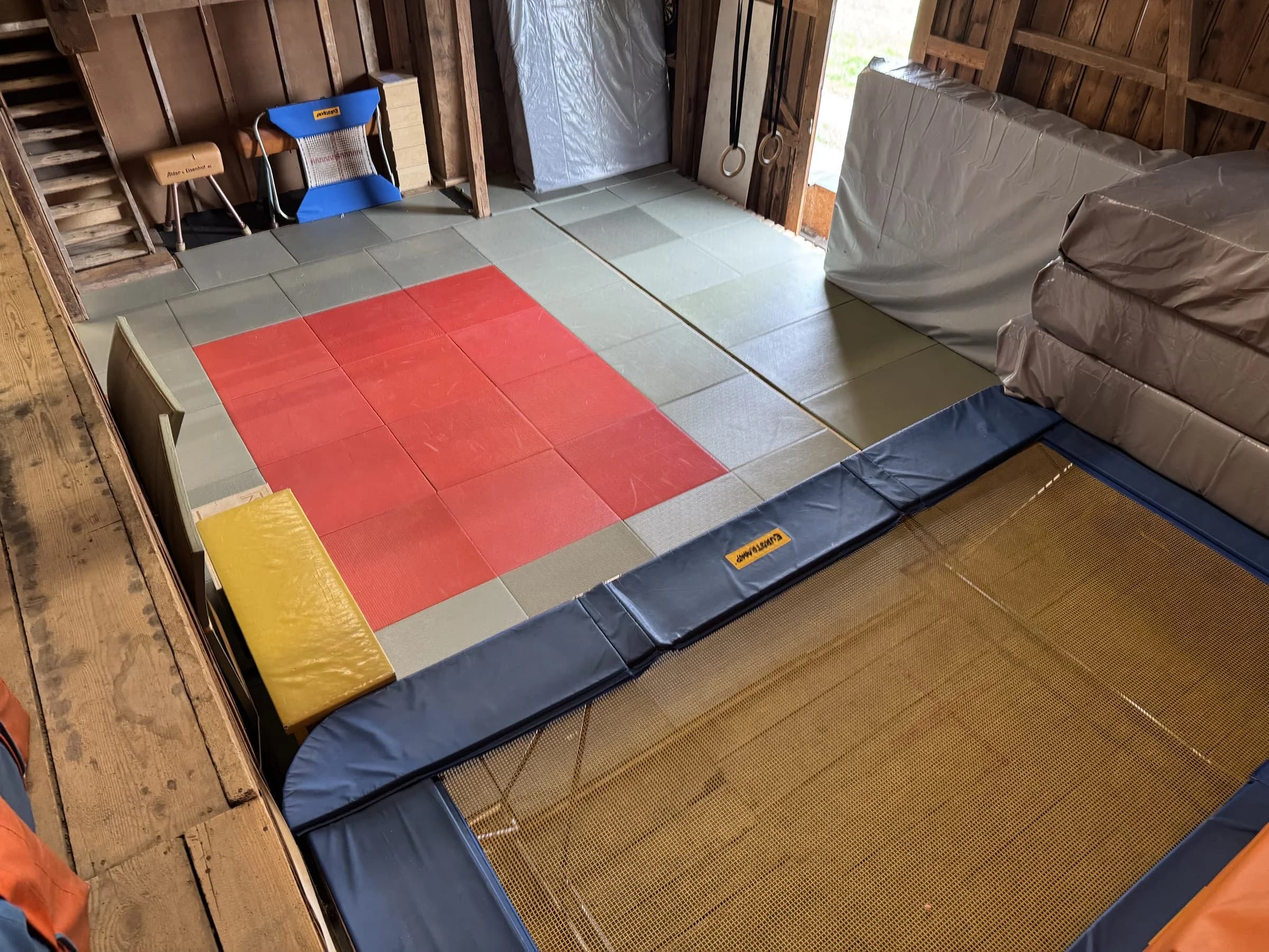 Erhöhte Mattenfläche mit rotem Feld, daneben das grosse Trampolin und gestapelte Weichböden