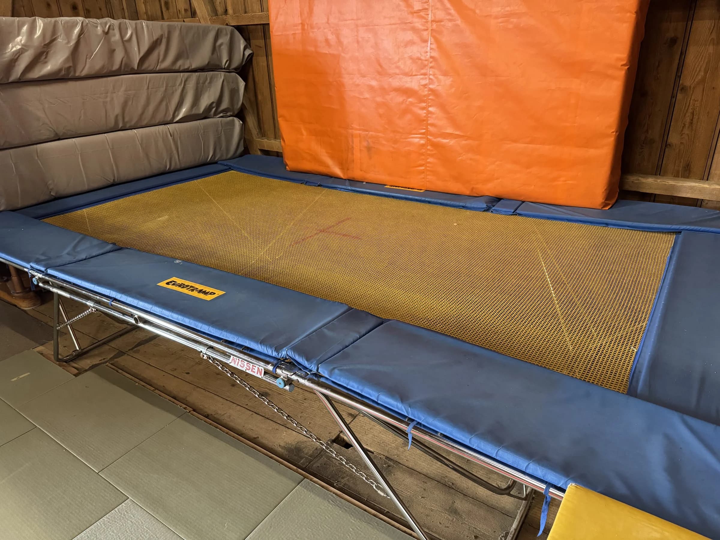 Grosses Trampolin mit blauen Randpolstern, daneben gestapelte Matten